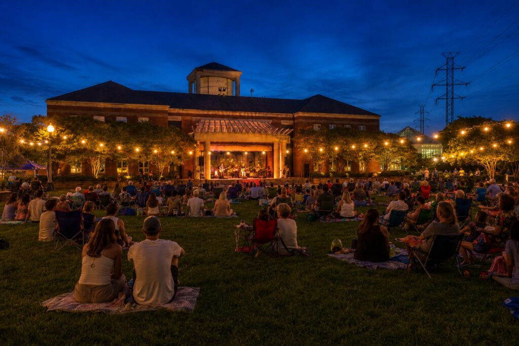 A free summer evening concert on the Herndon Town Green, with a crowd seated on the lawn under string lights